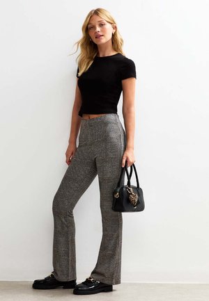 Vrouw in zwarte cropped t-shirt, geruite high-waisted flared broek, zwarte loafers, en met een zwart handtasje met een luipaard hart charm.