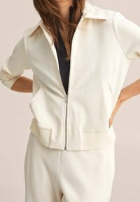 Chaqueta blanca con cremallera y cuello puntiagudo, bolsillos laterales y puños acanalados. La tela parece suave y lisa con un ajuste relajado.