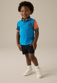 Turkos polo skjorta med kontrasterande orange ärmar, marinblå shorts, vita sneakers och textil med struktur. Har en logotyp på bröstet.