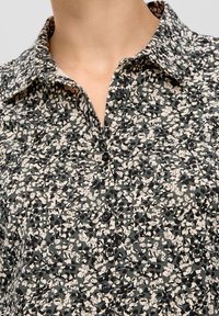 Button-up shirt with a collared neckline in a green, black, and cream abstract pattern. Features black buttons and a textured fabric.
