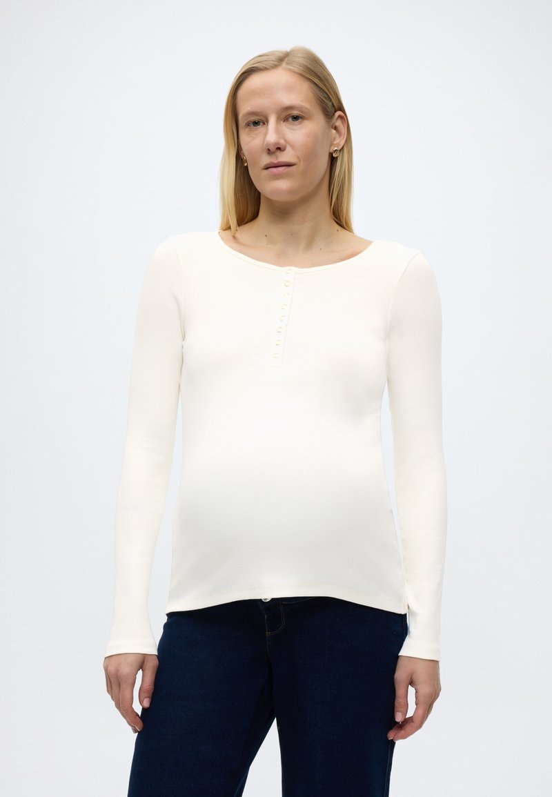 Έγκυος γυναίκα με μακριά ξανθά μαλλιά, φορώντας λευκό πουκάμισο μακρυμάνικο και σκούρο μπλε τζιν, μπροστά από ένα απλό φωτεινό φόντο.