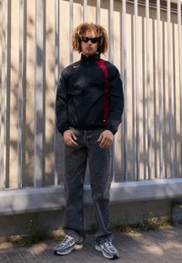 Veste noire avec des accents rouges, jean gris et baskets. Tissu texturé, fermeture à glissière et détail du logo sur la manche. Lunettes de soleil portées.