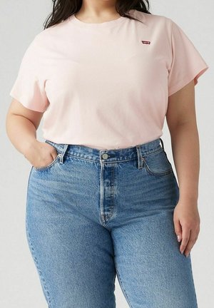 Woman wearing light pink Levi's t-shirt tucked into high-waisted blue jeans with one hand in pocket against a neutral background.