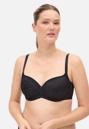 Mujer de piel clara y cabello castaño recogido en una coleta baja, vistiendo un sujetador negro de encaje con aros, mirando ligeramente hacia la derecha sobre un fondo claro y liso.