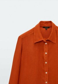 Chemise en lin couleur rouille avec un col classique, des manches longues et des boutons blancs sur le devant pour la fermeture.