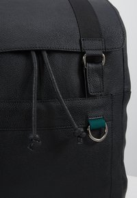 Svart texturerad läderväska med klaffstängning, dragsko-detalj och en silverring med en grön tygdetalj.