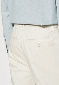 Maglione corto azzurro chiaro sopra pantaloni beige a vita elastica con dettagli sulla tasca posteriore e accentuazione con bottone. Texture del tessuto liscia.