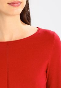 Femme portant un haut en maille rouge à encolure ronde, montrant une partie de son visage, son cou et son épaule sur un fond uni.