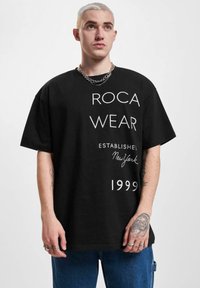 Melna lielizmēra T-kreksa ar baltu grafiskā teksta uzrakstu "ROCA WEAR ESTABLISHED New York 1999." Asistē īsas piedurknes un brīvu piegriezumu.