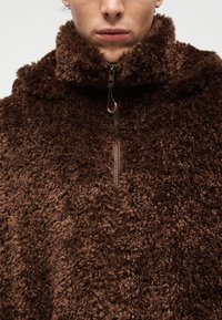 Καφέ φλις πουλόβερ με ψηλό γιακά και φερμουάρ. Το ύφασμα έχει χνουδωτή υφή, με μαλακή, πυκνή υφή παντού.