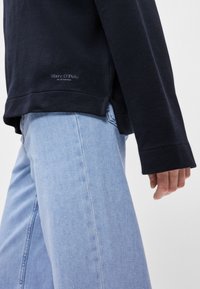 Sweatshirt azul-marinho de manga longa com fendas laterais, combinado com jeans azul-claro. O logótipo é apresentado de forma subtil perto da bainha do sweatshirt.