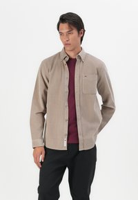Brown corduroy button-up shirt with a chest pocket, layered over a burgundy tee. The shirt features a collar and a subtle logo tag at the hem.