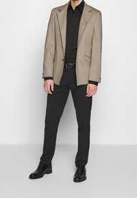 Beige skräddarsydd kavaj över en svart skjorta, i kombination med svarta byxor och svarta läderskor. Kavajen har två framfickor.