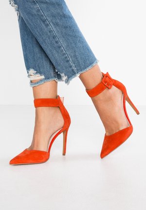 Ben i beskårne, frynsede blå jeans iført lyse orange ruskindspidsede høje hæle med ankelstropper og spænder.