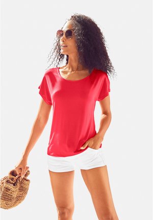 Rotes kurzärmliges Oberteil aus weichem Stoff, runder Ausschnitt, kombiniert mit weißen Shorts und einer gewebten Tasche. Modell trägt übergroße Sonnenbrille.
