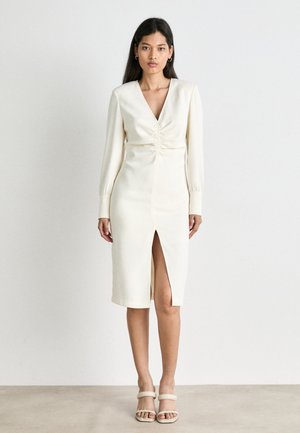 Cream-colored dress with a V-neck, long sleeves, ruched bodice, and a front slit. Smooth texture, knee-length silhouette, paired with neutral sandals.