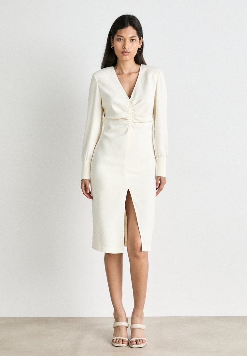 Cremefarbenes Kleid mit V-Ausschnitt, langen Ärmeln, gerafftem Oberteil und einem Schlitz vorne. Glatte Textur, knieumspielende Silhouette, kombiniert mit neutralen Sandalen.