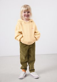 Jeune enfant souriant, portant un sweat à capuche jaune, un pantalon en velours côtelé vert olive, des chaussettes orange et des baskets blanches, debout devant un arrière-plan uni.