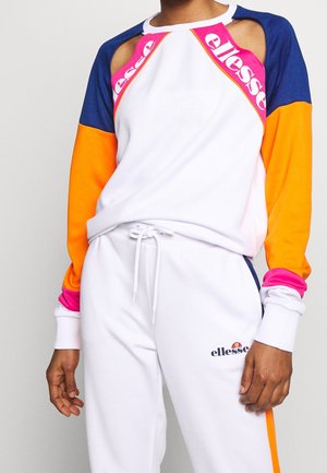 Osoba ubrana w białe spodnie i pasującą białą bluzę z niebieskimi, pomarańczowymi i różowymi rękawami, z logo „ellesse” na piersi i nodze.