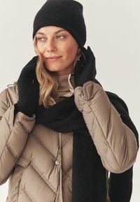 Hengittävä beige puhujatakki, jossa on timanttinilkkinen kuvio, yhdistetty mustaan neulehuiviin ja pipoon. Mustat hanskat ja korkea kauluksen vaaleanruskea toppi näkyvät.