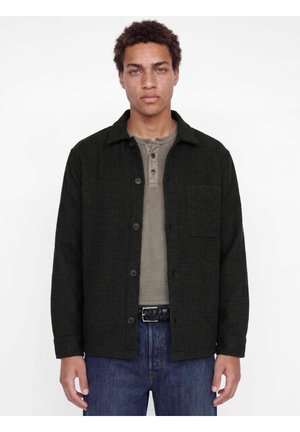 Jeune homme aux cheveux bouclés porte une veste-chemise sombre boutonnée sur un t-shirt henley beige et un jean bleu sur un fond blanc uni.