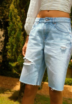 Shorts de mezclilla azul claro tipo bermuda con parches desgastados, dobladillo deshilachado y diseño clásico de cinco bolsillos. Presenta herrajes en tono cobre.
