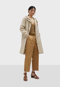 Lekki beżowy płaszcz przeciwdeszczowy w białe kropki, brązowa warstwowa bluzka i beżowe spodnie; uzupełniony brązowymi sandałami na paski i wyrazistymi kolczykami.