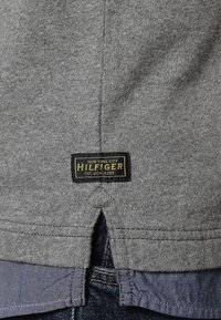 Grau Hoodie mit split Hemd hinten, versehen mit einem schwarzen Etikett mit gelber Schrift, auf dem "HILFIGER" und "NEW YORK CITY" steht. Weicher Stoff.