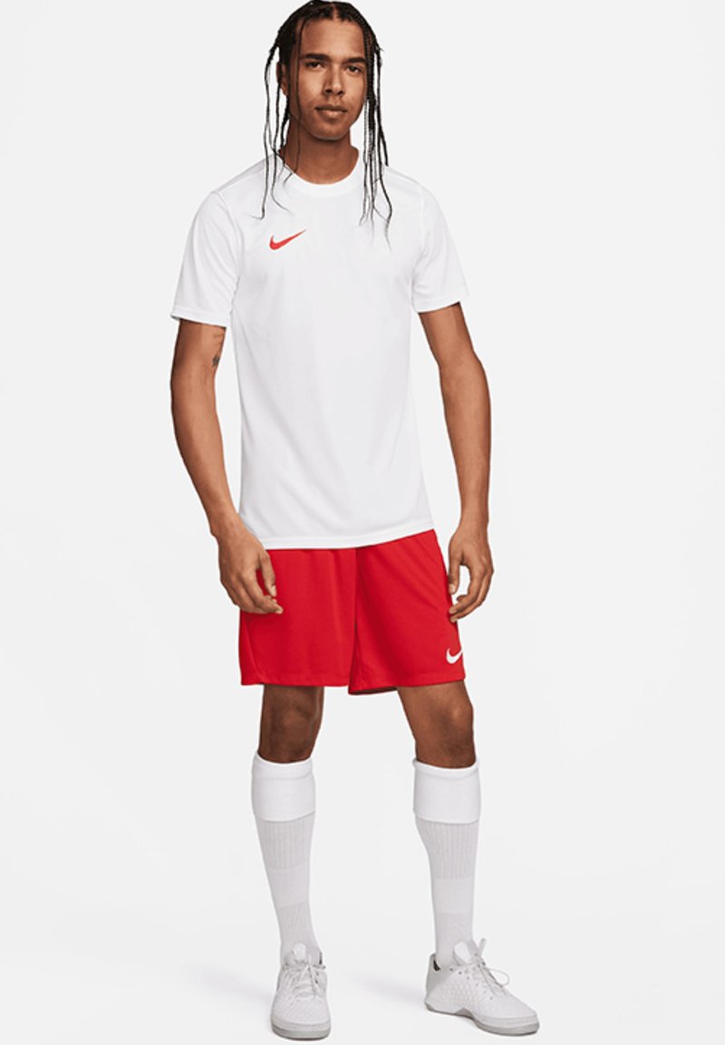 Maglietta Nike bianca con logo rosso, pantaloni corti sportivi rossi, calzini bianchi al ginocchio e sneakers grigie, con un fit casual e tessuto traspirante.