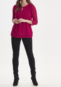 Blusa magenta con scollatura a chiave e maniche a tre quarti, abbinata a pantaloni neri aderenti e stivaletti neri. Texture liscia.