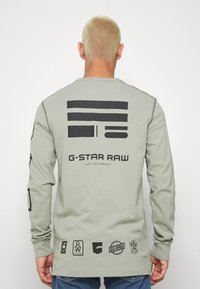 Langärmliges olivgrünes Hemd mit schwarzen geometrischen Grafikdesigns und Logos auf der Rückseite, mit gerippten Bündchen und Rundhalsausschnitt.