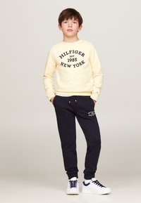 Felpa gialla con stampa nera "Hilfiger New York", abbinata a pantaloni da jogging blu navy con logo bianco e sneakers bianche con dettagli blu navy.