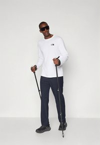 Biela košeľa s dlhým rukávom s logom, tmavomodré nohavice a čierne turistické topánky. Drží trekingové palice a má na sebe tmavé slnečné okuliare.