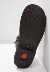 Semelle de chaussure en caoutchouc noir avec surface texturée, comportant un accent de feuille rouge et un logo "Kickers" en relief. Forme arrondie avec durabilité.
