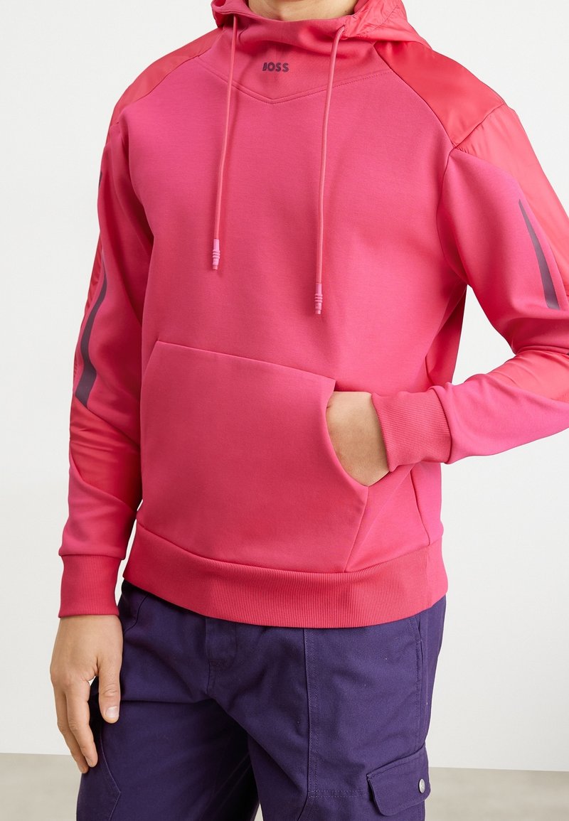 Różowa bluza z kapturem, posiadająca kieszeń kangurzą, sznurki oraz logo na klatce piersiowej. Charakteryzuje się kontrastującymi rękawami z szarymi akcentami. Gładka tekstura.