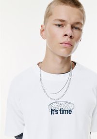 Ung mand med kort lyst hår iført hvid T-shirt med teksten "It's time" og to sølv kæder, foran en ensfarvet hvid baggrund.
