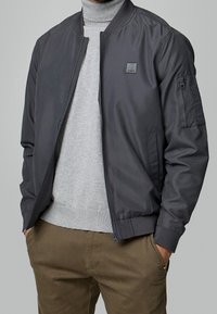 Donkergrijze bomberjack van soepel materiaal, met een rits aan de voorkant, geribbelde kraag en mouwzakdetails. Draag het over een lichtgrijze trui.