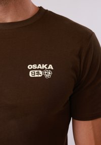 Gros plan sur un t-shirt marron porté par une personne, avec le texte blanc « OSAKA » accompagné de caractères japonais et d'un motif floral sur la poitrine.