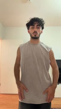 Sleeveless light gray tank top with a round neckline, smooth texture, and slightly dropped armholes. Room interior in background.