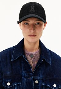 Junge Person mit Sommersprossen, die eine schwarze Cordmütze, eine dunkelblaue Cordjacke und einen lila Strickpullover trägt und mit dem Gesicht zu einer weißen Hintergrund steht.