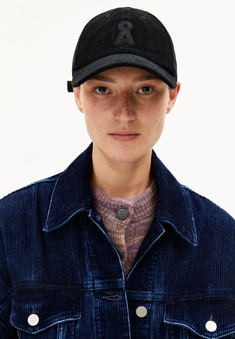 Junge Person mit Sommersprossen, die eine schwarze Cordmütze, eine dunkelblaue Cordjacke und einen lila Strickpullover trägt und mit dem Gesicht zu einer weißen Hintergrund steht.