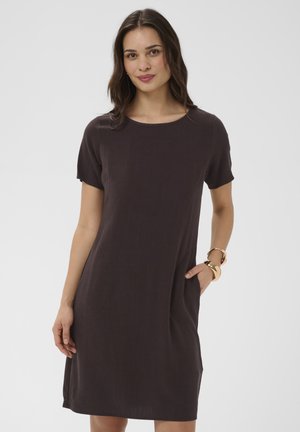 Femme debout, main dans la poche, portant une robe marron foncé à manches courtes arrivant au genou et des bracelets dorés sur un fond blanc.