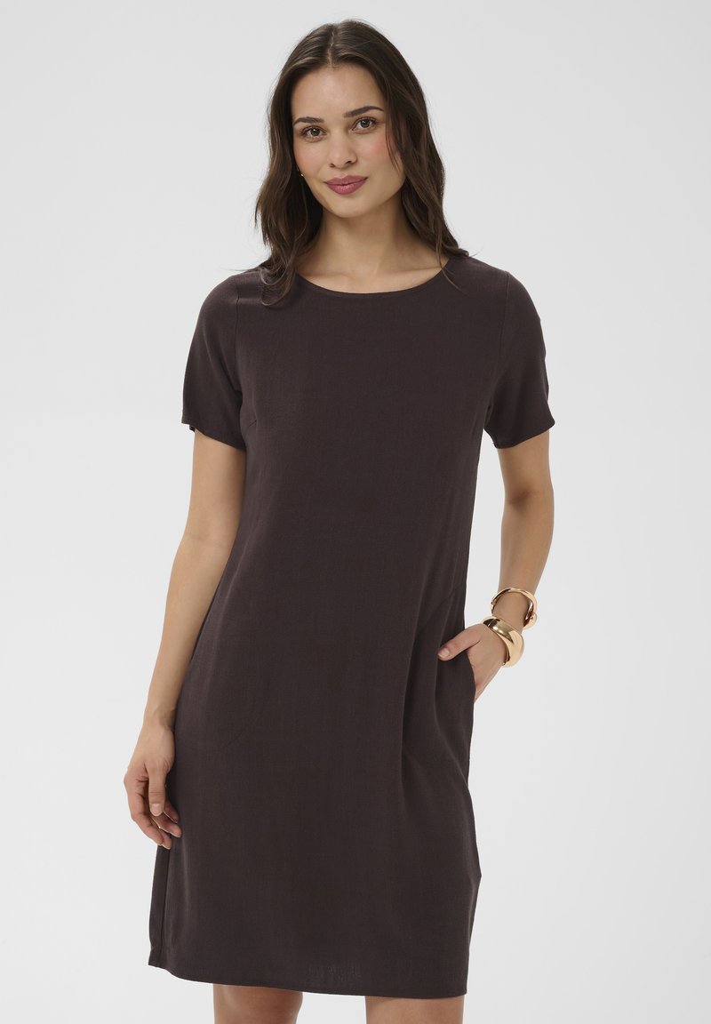 Femme debout, main dans la poche, portant une robe marron foncé à manches courtes arrivant au genou et des bracelets dorés sur un fond blanc.