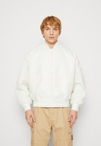 Chaqueta bomber blanca con cremallera frontal, cuello y puños acanalados, y cinturilla elástica. Textura suave con un ligero brillo. Llevada sobre pantalones cargo beige.