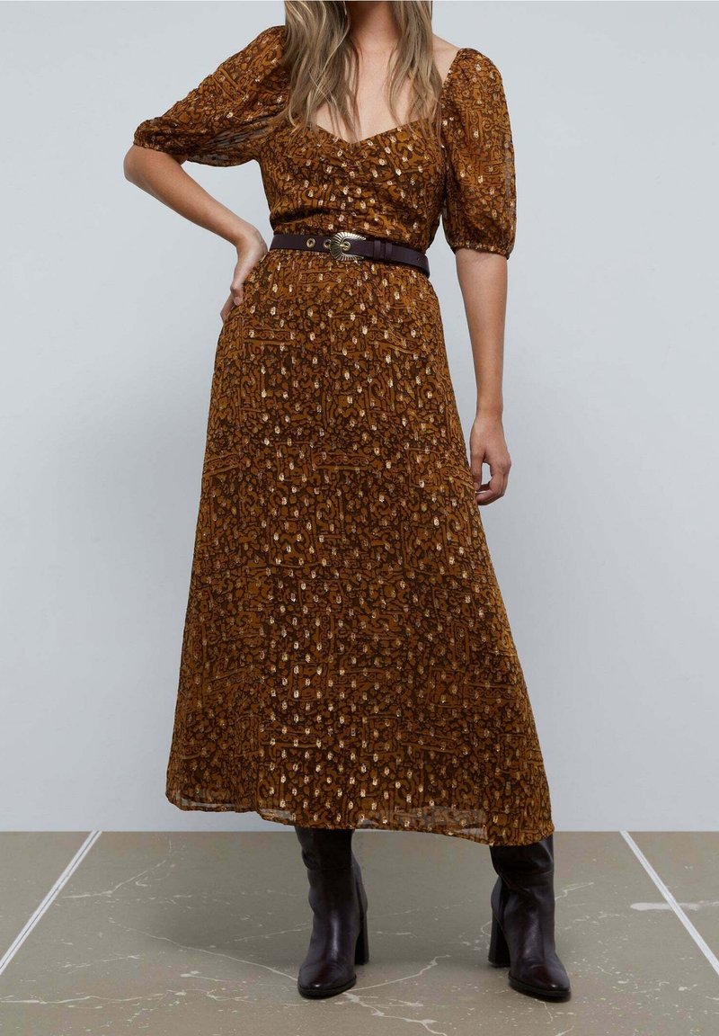 Robe maxi marron à motifs avec manches bouffantes, encolure en V et taille cintrée agrémentée d'une ceinture marron foncé. Bottines noires à talons complètent le look.