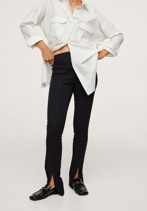 Kobieta ubrana w czarne obcisłe spodnie z rozcięciami z przodu na nogawkach, czarne mokasyny oraz luźną białą koszulę z podwiniętymi rękawami.