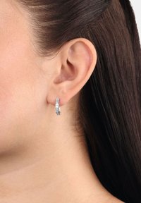 Primo piano di un orecchio di donna con capelli scuri lisci che indossa un piccolo orecchino a cerchio in argento con design texturizzato.