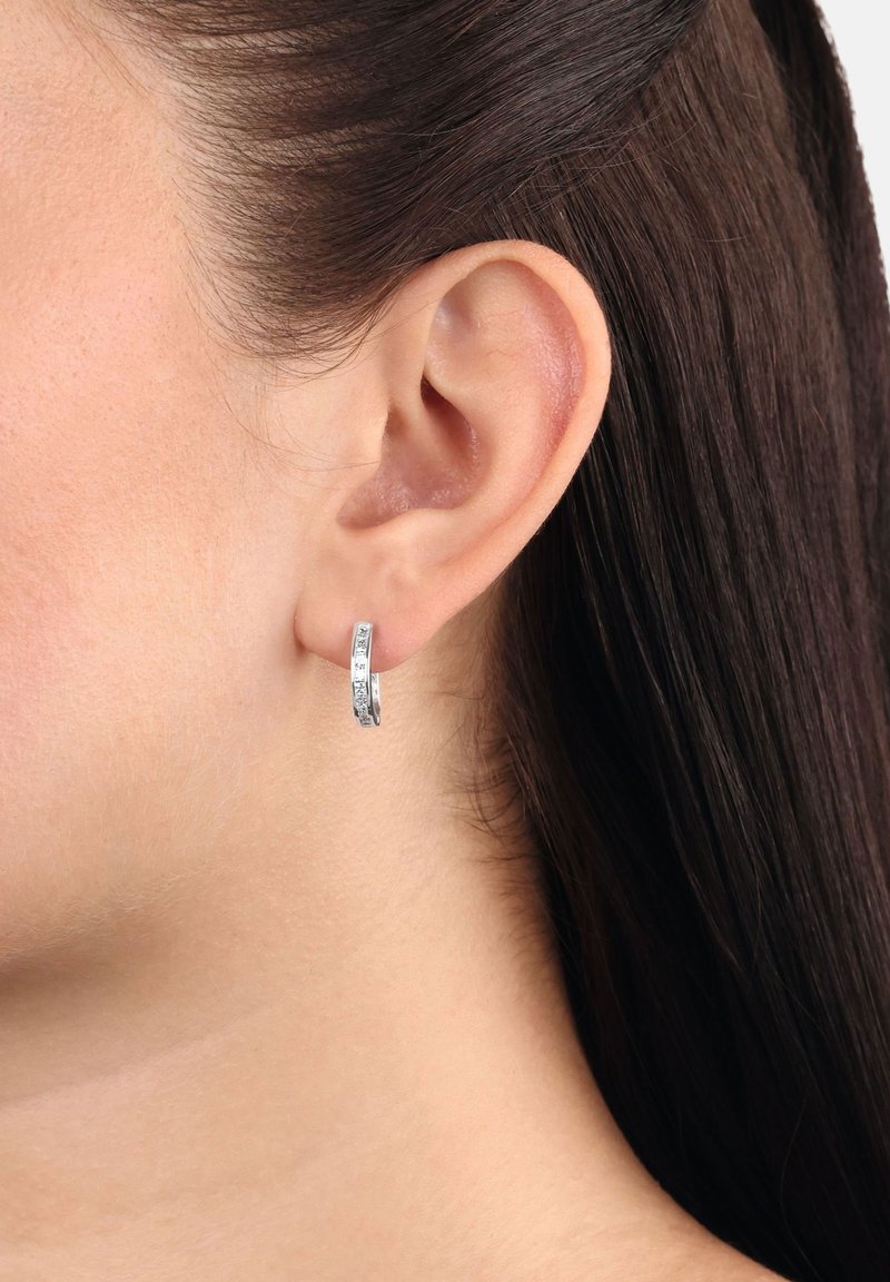 Primo piano di un orecchio di donna con capelli scuri lisci che indossa un piccolo orecchino a cerchio in argento con design texturizzato.