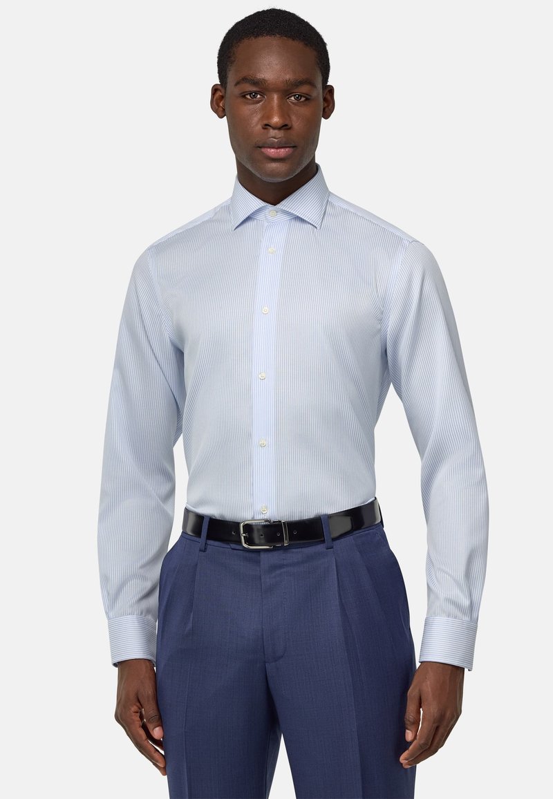 Camisa de vestir a rayas en azul claro con cuello, cierre frontal de botones y mangas largas, combinada con pantalones oscuros de corte ajustado y un cinturón negro.