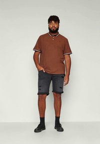 Homem em pé com uma mão no bolso, vestindo uma t-shirt polo castanha, calções de ganga pretos rasgados, meias pretas e sapatilhas pretas.